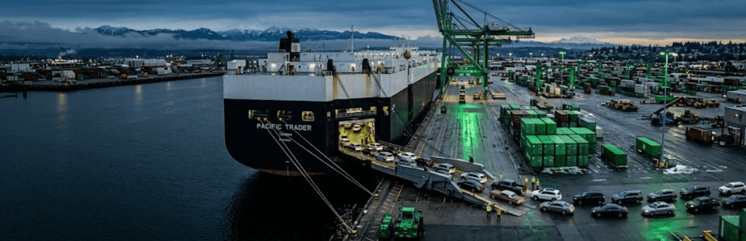 RoRo car carrier ship unloading vehicles down a ramp at a port terminal at dusk, with green cranes and stacked containers