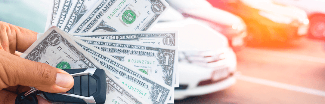 Hand holding a fan of dollar bills and a car key in front of a row of cars