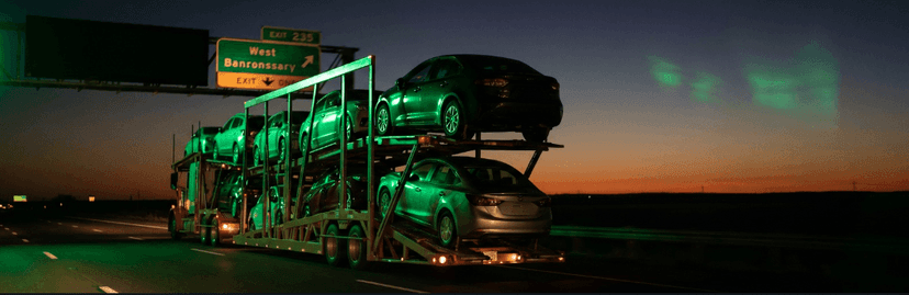 Car carrier lit in green on a highway at dusk under a West sign heading toward California