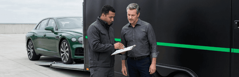 Two men reviewing a document beside a dark green Bentley sedan and an enclosed trailer