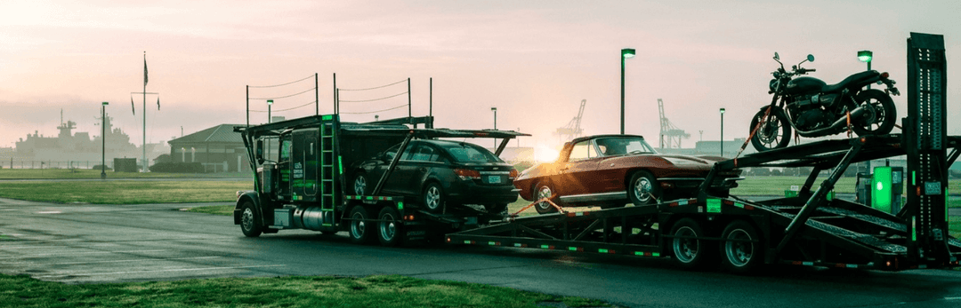 Car carrier loaded with a classic car, a sedan and a motorcycle parked by a field at dawn