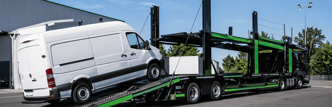 White cargo van driving up the ramp of a green open car carrier trailer in a parking lot