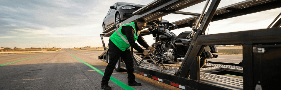 Worker in a green safety vest strapping a motorcycle onto the deck of an open car carrier trailer
