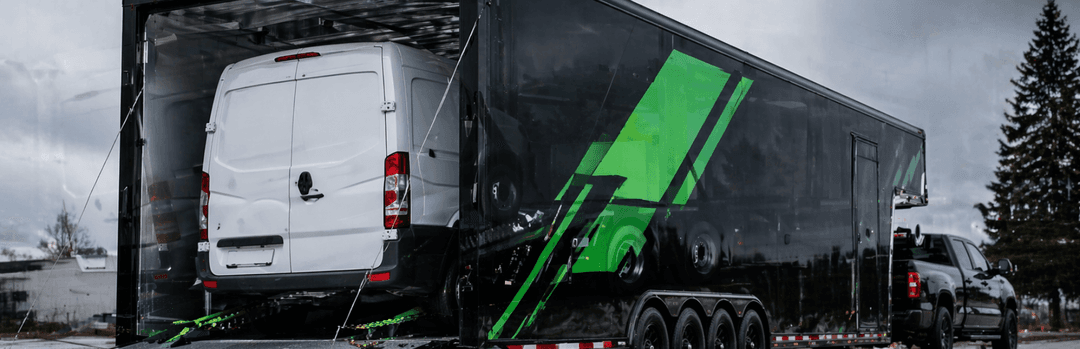 White cargo van loaded inside a black enclosed transport trailer with a green stripe on a rainy day
