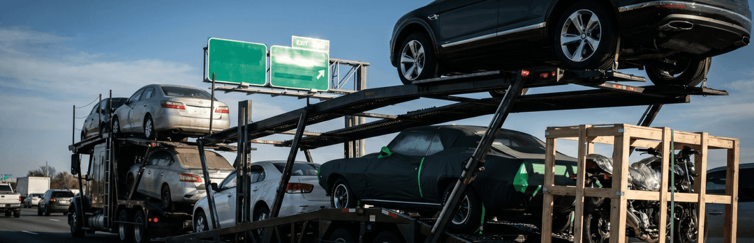 Open car carrier loaded with sedans beside a wooden crate under green highway signs